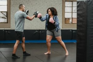 Ethnic male instructor with focus mitts combating with overweight African American female athlete while practicing boxing in gymnasium