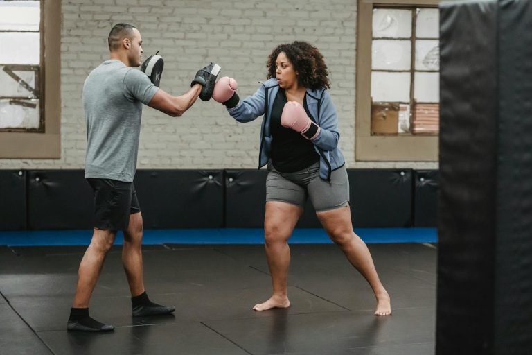 Ethnic male instructor with focus mitts combating with overweight African American female athlete while practicing boxing in gymnasium